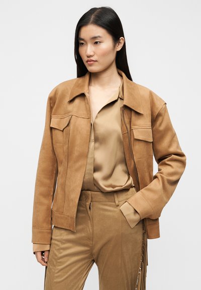 Μπεζ καφέ σαμπρέ σακάκι με γιακά, τσέπες στο στήθος και άνετη εφαρμογή. Φορεμένο με αντίστοιχο μπεζ μπλούζα και παντελόνια.