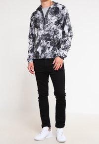 Bluza z kapturem w abstrakcyjny wzór czarno-biały, lekka tkanina, z przodem na półzamek, kieszenią typu kangur, zestawiona z czarnymi spodniami i białymi trampkami.