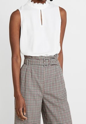 Blouse blanche sans manches avec col en dentelle et encolure en trou de serrure, associée à un pantalon à carreaux taille haute avec ceinture.