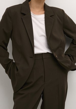 Personne portant un costume marron foncé sur mesure avec blazer et pantalon taille haute assorti, par-dessus un t-shirt blanc uni à col rond.