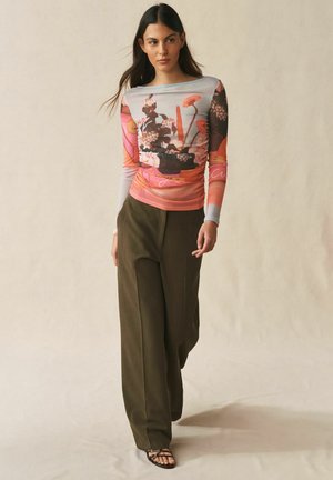 Ung kvinde går, iført en langærmet blomstret top, olivengrønne bukser med vide ben og sorte åbne sandaler på en neutral baggrund.