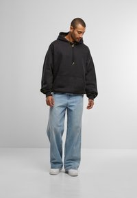 Schwarzer Kapuzenpullover mit Fronttasche, goldene Aglets an den Kordeln, kombiniert mit hellblauen weit geschnittenen Jeans und weißen Sneakers.