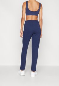 Henkilö, joka on pukeutunut tummansinisiin verkkohousuihin, tummansiniiseen urheiluliiviin ja valkoisiin tennareihin, seisoo selkä kameraan päin yksinkertaista taustaa vasten.