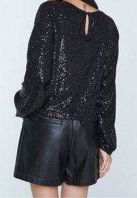 Top nero con paillettes e maniche lunghe, caratterizzato da un dettaglio a chiave sul retro. Abbinato a pantaloni corti in pelle nera a vita alta. Texture liscia.