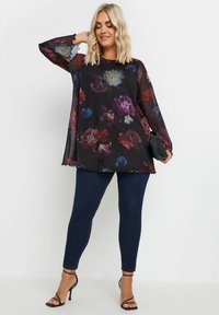 Top trasparente nero floreale con maniche lunghe e orlo a scallop, abbinato a leggins blu scuro e sandali con tacco neri.
