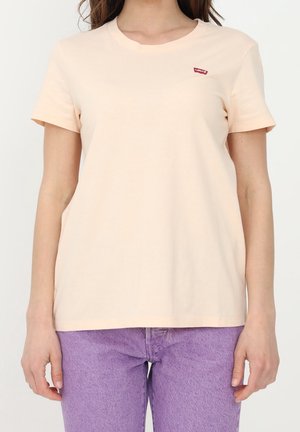Femme portant un t-shirt simple beige clair à manches courtes avec un petit logo rouge Levi’s sur la poitrine et un jean en denim violet.