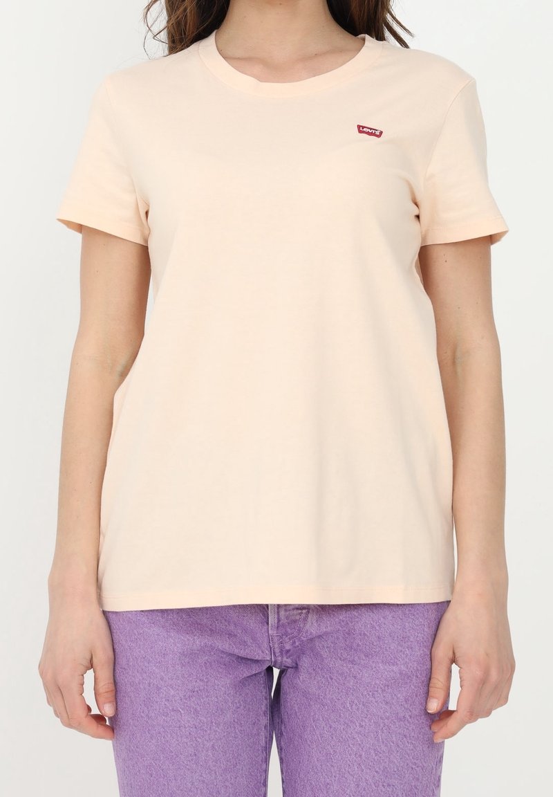 Femme portant un t-shirt simple beige clair à manches courtes avec un petit logo rouge Levi’s sur la poitrine et un jean en denim violet.