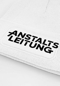 White knitted beanie with a textured pattern, featuring a rectangular patch with black lettering "ANSTALT'S LEITUNG" and a stitched border.