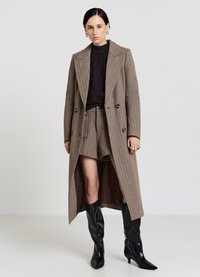 Cappotto lungo a righe marroni con bottoni doppiopetto, indossato sopra un maglione scuro e pantaloncini abbinati, abbinato a stivali neri al ginocchio.