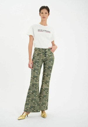 Pantalons large à motifs en vert et beige avec divers motifs géométriques et botaniques ; associés à un t-shirt blanc avec logo et des chaussures dorées.