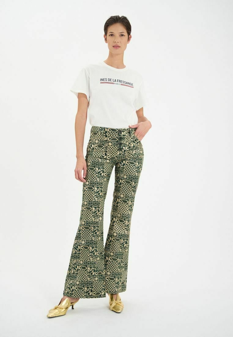 Pantalons large à motifs en vert et beige avec divers motifs géométriques et botaniques ; associés à un t-shirt blanc avec logo et des chaussures dorées.
