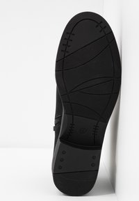Semelle de chaussure noire avec motifs de traction texturés et marquage de taille 37, inclinée contre un fond blanc d'angle.