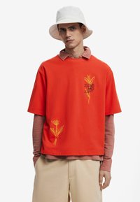 Rød kortærmet bomulds T-shirt med gul blomsterbroderi, båret over en lyserød langærmet T-shirt, kombineret med beige bukser.