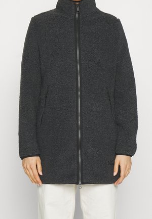 Person trägt eine lange schwarze, strukturierte Fleecejacke mit Vorderreißverschluss und seitlichen Reißverschlusstaschen, kombiniert mit weißen Hosen.