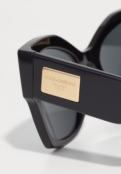 Ochelari de soare negri cu un cadru geometric, având lentile închise la culoare și o plăcuță din metal aurit cu logo-ul care spune "DOLCE & GABBANA MILANO ITALIA."