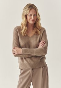 Pruun kudumipullover V-kaelusega, pikkade varrukatega ja sooniku äärtega. Kangas näeb v�älja pehme ja tekstuurne. Koos beežide pükstega.