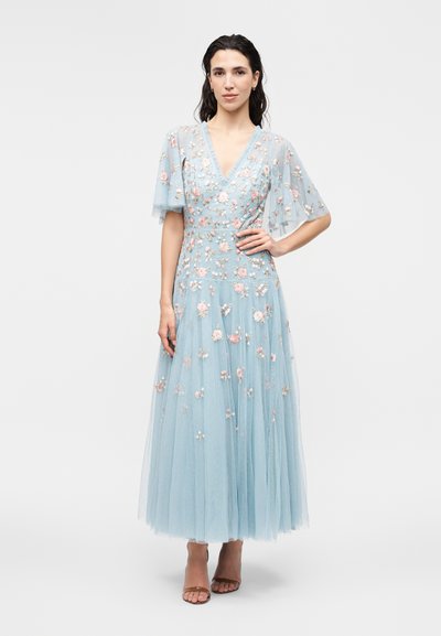 Kvinde iført en lyseblå, gulvlængde kjole med gennemsigtige flagrende ærmer og pastel blomsterbroderi, stående med den ene hånd på hoften.
