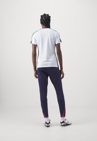 Conjunto deportivo que incluye una camiseta blanca de manga corta con detalles en negro y pantalones morados con adornos en rosa, combinados con calzado en blanco y negro.