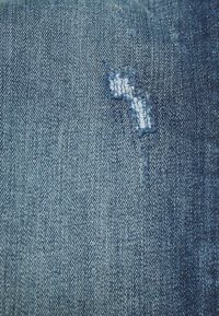 O tecido de denim apresenta uma cor azul clara com uma área desgastada mostrando bordas desfiadas. A textura é suave, com padrões de trama visíveis.