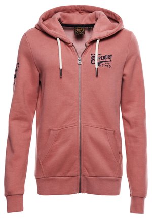 Prachově růžová mikina na zip s bílými šňůrkami, předními kapsami, žebrovanými manžetami a černým logem „Superdry“ na hrudi a levém rukávu.