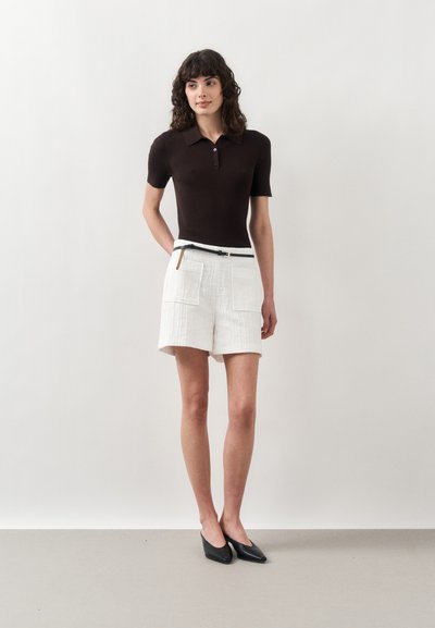 Femme aux cheveux bouclés portant un polo noir à manches courtes, un short blanc ceinturé avec des poches, et des chaussures noires à bout pointu, debout sur un fond blanc.