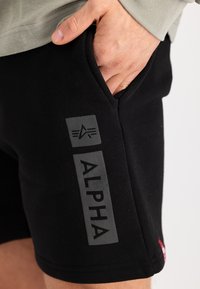 Mustat urheilushortsit, joissa on harmaa "ALPHA" -logo sivussa ja pieni tasku. Valmistettu pehmeästä, joustavasta kankaasta, jossa on sileä pinta.