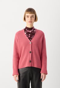 Rožinė, raukšlėta megztinė su V formos iškirpte ir trimis rudos spalvos mygtukais, su ilgomis rankovėmis ir laisvu siluetu, derinama su raštuota polo kaklo maike.