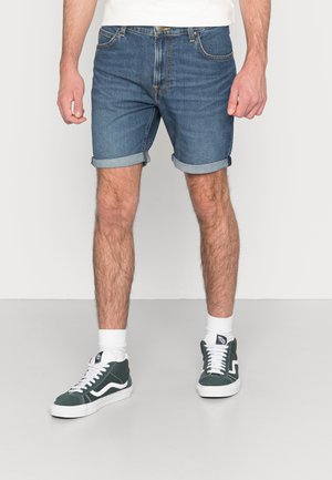 Mand iført op rullede blå cowboyshorts, hvide sokker og grønne og hvide sneakers, stående på en grå gulv.