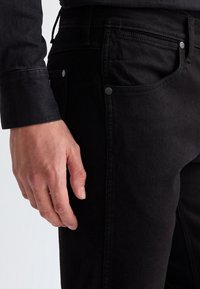 Mâna stângă odihnește pe lângă corp, purtând o cămașă neagră cu mânecă lungă și blugi negri cu buzunar frontal vizibil și nituri metalice.