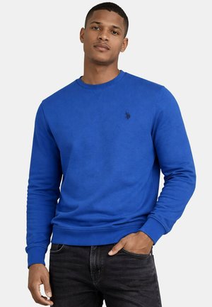 Mann trägt ein blaues Sweatshirt mit Rundhalsausschnitt und kleinem dunklem Logo auf der linken Brust, eine Hand in der Tasche seiner schwarzen Jeans, schaut nach vorne.