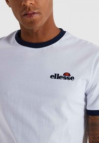 Valge puuvillane T-särk tumesinise soonikkraega ja varrukate äärtega. Omab tikandiga "ellesse" logo, millel on punane ja must aksent.
