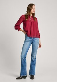 Blusa trasparente bordeaux con collo a fiocco, maniche lunghe e polsini arricciati, abbinata a jeans svasati azzurri chiari e stivaletti neri con tacco.