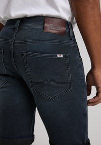 Jeans de mezclilla oscura con una etiqueta de cuero marrón en la cintura. Diseño distintivo en el bolsillo trasero con una pequeña etiqueta de logo. Puños enrollados.