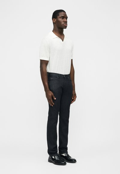 Άνδρας όρθιος, φοράει λευκό πόλο με κοντά μανίκια, μαύρα τζιν και μαύρα δερμάτινα μοκασίνια, μπροστά σε απλό ανοιχτό φόντο.