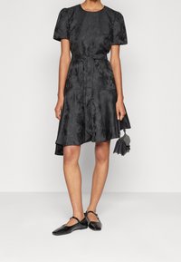 Robe noire avec des manches bouffantes courtes, tissu texturé, détail de ceinture à la taille et ourlet asymétrique. Accompagnée de chaussures plates noires et d'un petit sac.