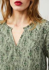 Grön mönstrad blus med svarta och gröna blommönster, med V-ringning och subtil veckning runt halslinningen. Slät tygtextur.