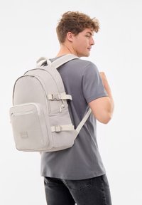 Sac à dos en tissu gris avec des bretelles réglables, une poche zippée à l'avant et une poche latérale texturée. Présente des accents de boucle et une forme arrondie.
