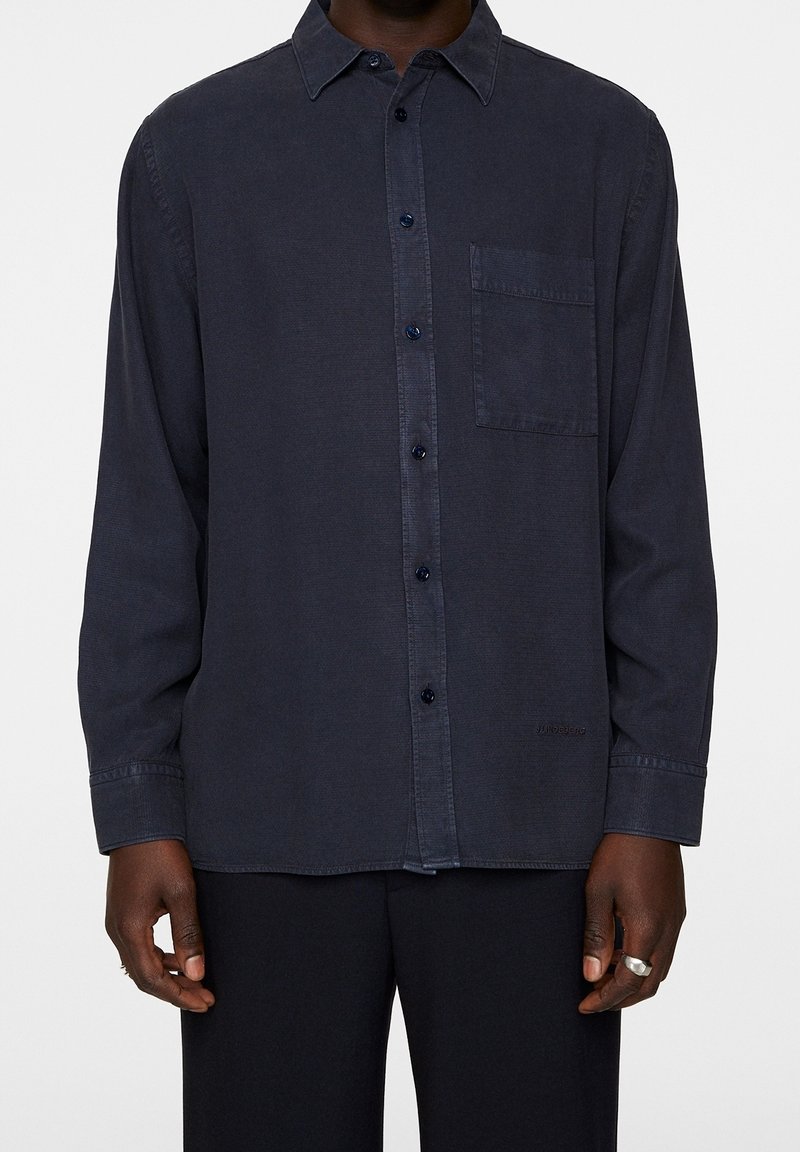 Homme portant une chemise bleu marine foncé à manches longues avec une poche poitrine et un pantalon noir, debout devant un fond uni.
