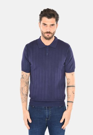 Uomo con capelli corti e scuri e barba, che indossa una polo blu navy a maniche corte e jeans blu, in piedi davanti a uno sfondo bianco.