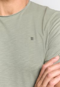 Lichtgroene T-shirt met korte mouwen, gemaakt van zachte stof, met een klein zwart logo op de linkerkant van de borst. Soepele textuur en ronde halslijn.