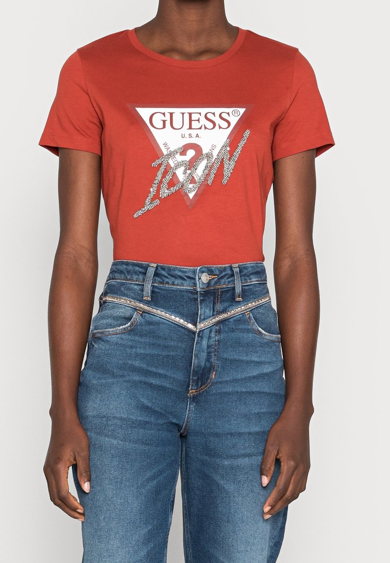 Personne portant un t-shirt rouge Guess Icon et un jean bleu taille haute avec une ceinture argentée ornée de strass.