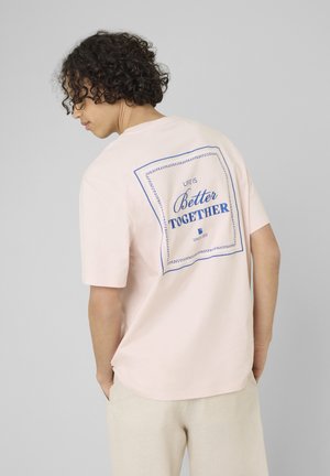 Personne aux cheveux bouclés portant un t-shirt rose pâle avec le texte bleu « Life is Better Together » dans le dos, debout devant un fond gris.