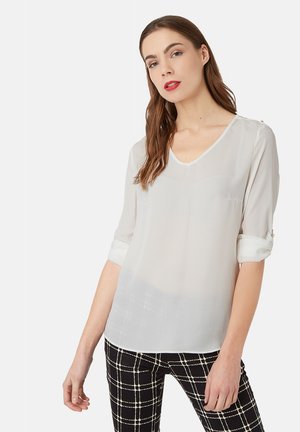 Mujer con largo cabello castaño lleva una blusa blanca transparente con mangas arremangadas y pantalones negros con un patrón de cuadros blancos.