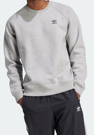 Mężczyzna w jasnoszarej bluzie Adidas i czarnych spodniach dresowych Adidas, z rękami w kieszeniach na jasnym tle.