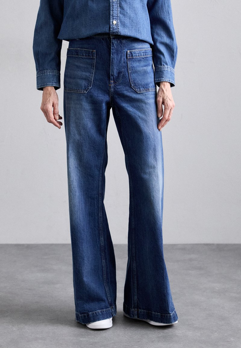 Flared blue denim jeans with a light wash, featuring front and back pockets, and contrast stitching. Worn with a blue denim shirt.