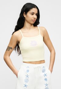 Νέα γυναίκα με μακριά σκούρα μαλλιά που φοράει ανοιχτόκίτρινο κοντό τοπ και λευκά σορτς με μπλε σχέδια θαλάσσιων πλασμάτων, τα χέρια στις τσέπες.