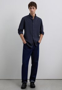 Camisa azul marino de cuadros con botones y mangas remangadas, combinada con jeans azul oscuro y zapatos negros, posando contra un fondo neutro.