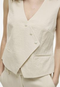 Beige ilmanhihaista liiviä, jossa on V-pääntie ja epäsymmetrinen muotoilu. Siinä on kolme yhteensopivaa nappia ja kaksi etutaskua teksturoitua kangasta.