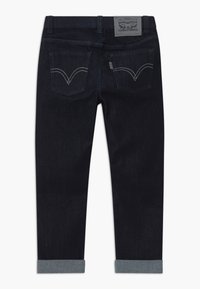 Dark denim jeans with a straight cut, featuring folded cuffs, two back pockets with a V-stitch pattern, and a logo patch on the waistband.