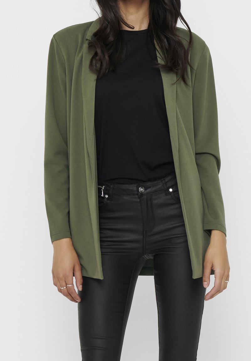 Femme portant une veste verte olive ouverte sur le devant, un chemisier noir et un pantalon en cuir noir, debout avec les mains détendues le long du corps.